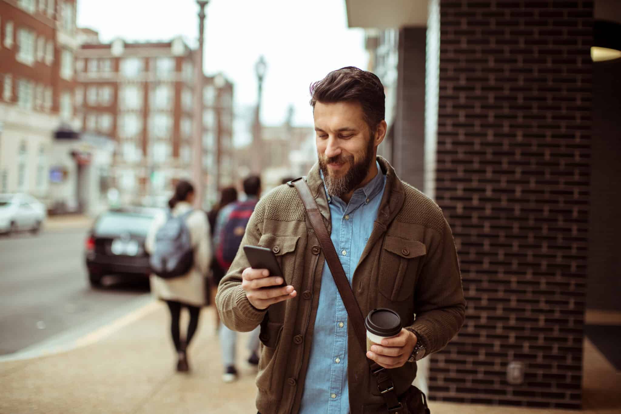 Man walking with coffee and using smartphone in city street reading about the rise of social media management tools